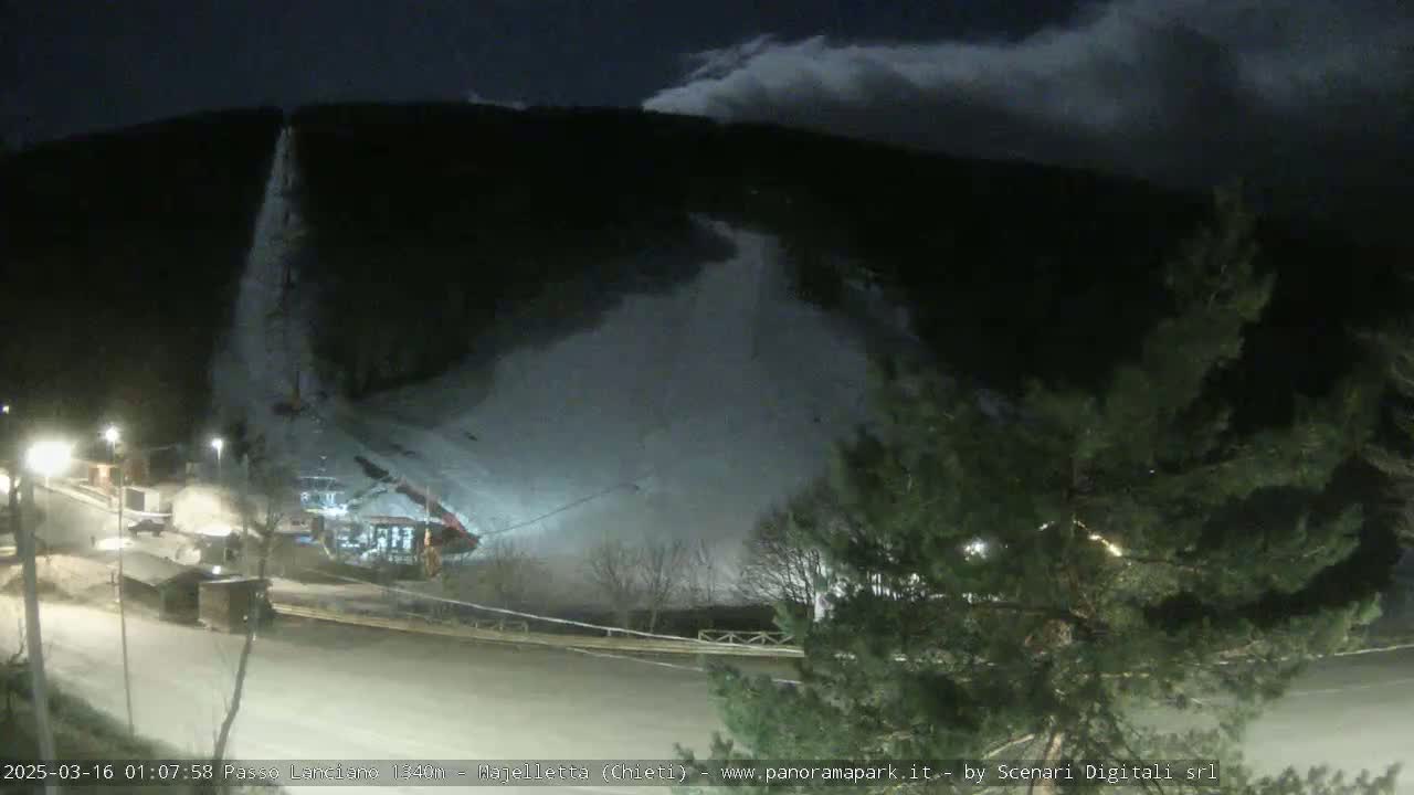 A snow-covered ski slope at night under a cloudy sky, with lights illuminating a base area.