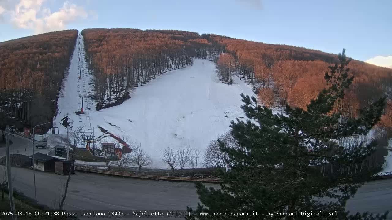 A snow-covered ski slope with a chairlift ascends a hillside of reddish-brown trees under a clear blue sky.