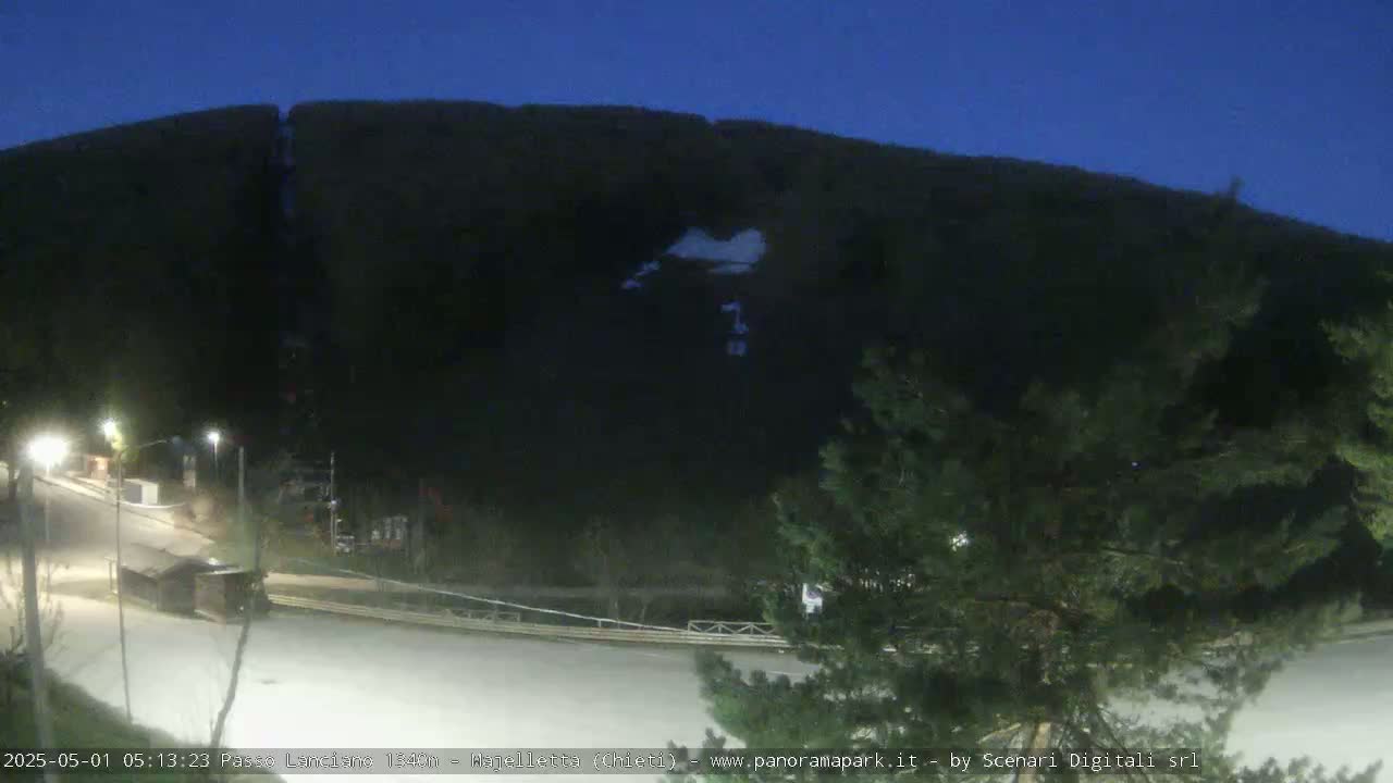 A nighttime view of a dark mountain with a small patch of snow, overlooking a mostly empty road and some sparse vegetation.