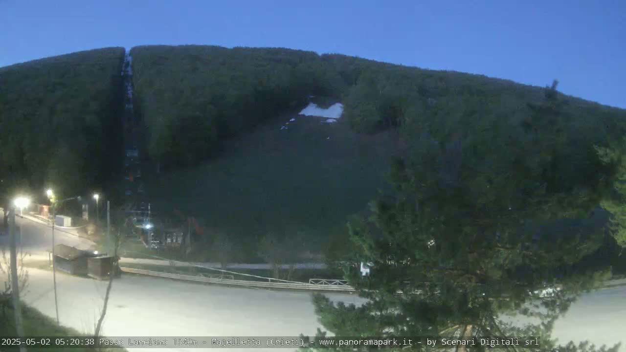 A nighttime, wide shot shows a mostly empty ski slope on a mountainside with a few small patches of snow remaining, under a clear, dark blue sky.