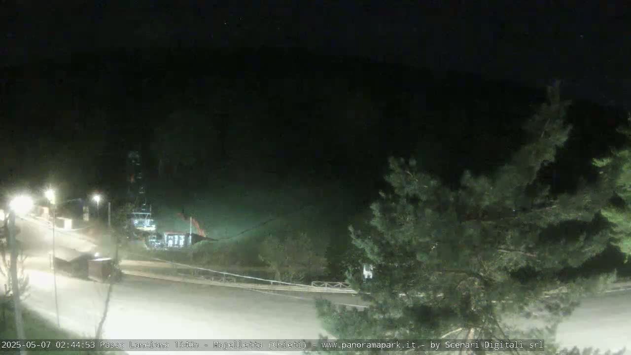 A nighttime view of a road curving through a dark, sparsely lit area with some trees and a structure in the background.