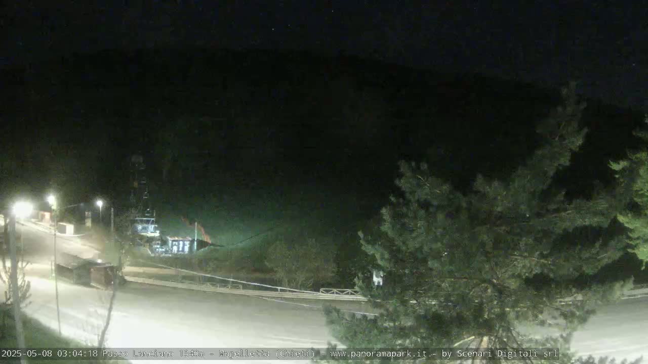 A nighttime, snow-covered road curves past a building and trees under a dark sky.