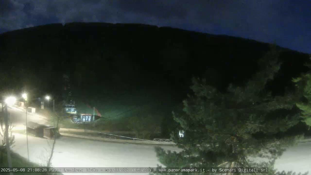 A nighttime, dimly lit outdoor scene shows a paved road curving around a dark mountainside with sparse vegetation and a few illuminated structures.
