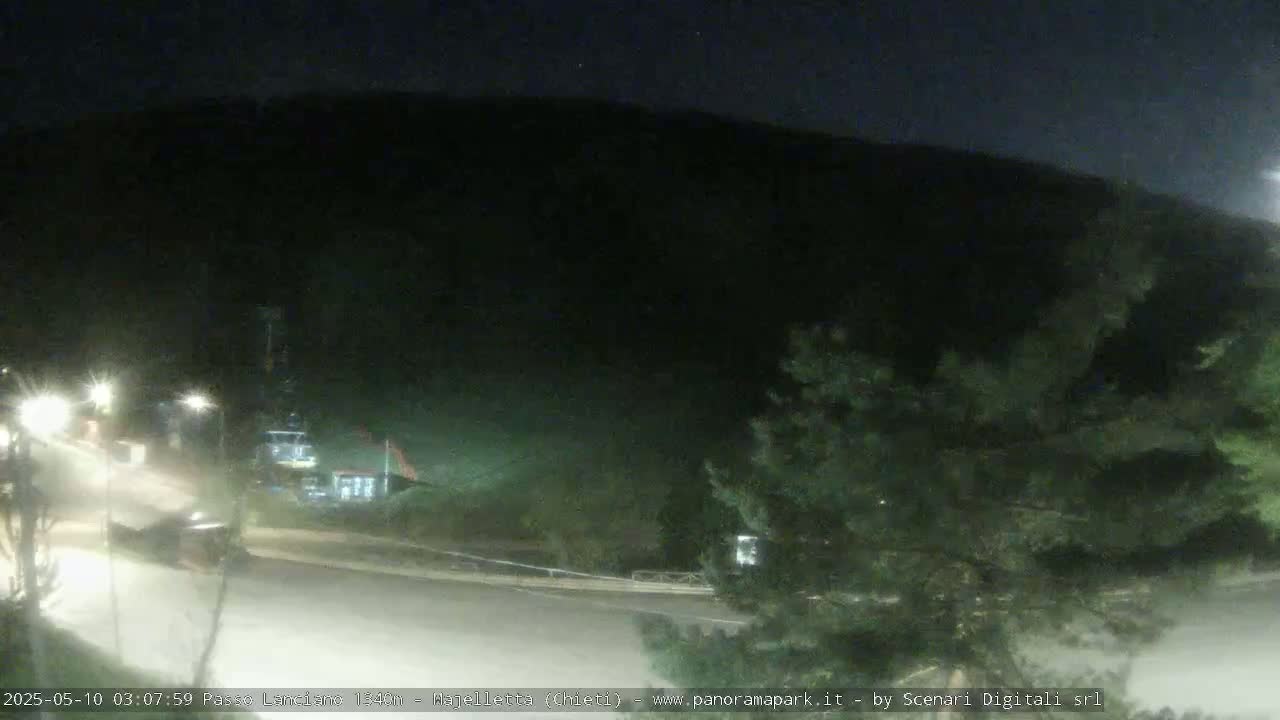 A nighttime, dimly lit road curves gently around a dark hillside, with a few vehicles visible and some sparse vegetation.