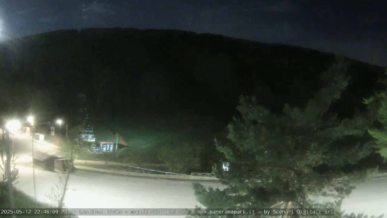 A nighttime, dimly lit view of a roadway curving toward a dark mountainside, with some trees and sparse vegetation visible in the foreground.