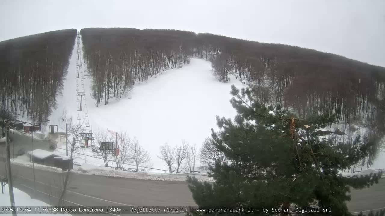 The image captures a snow-covered ski slope with a ski lift ascending between dense forests, a road, and small buildings visible in the foreground, all under an overcast sky.