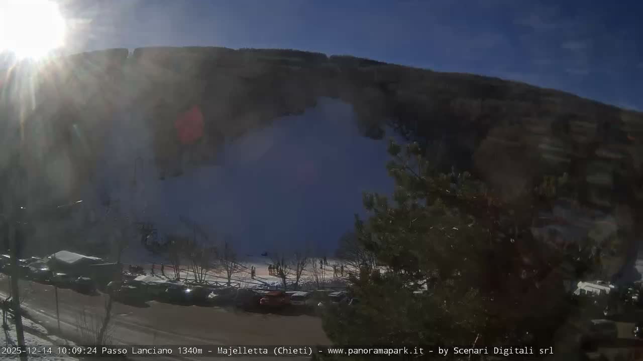 The image captures a snow-covered ski slope with a ski lift ascending between dense forests, a road, and small buildings visible in the foreground, all under an overcast sky.