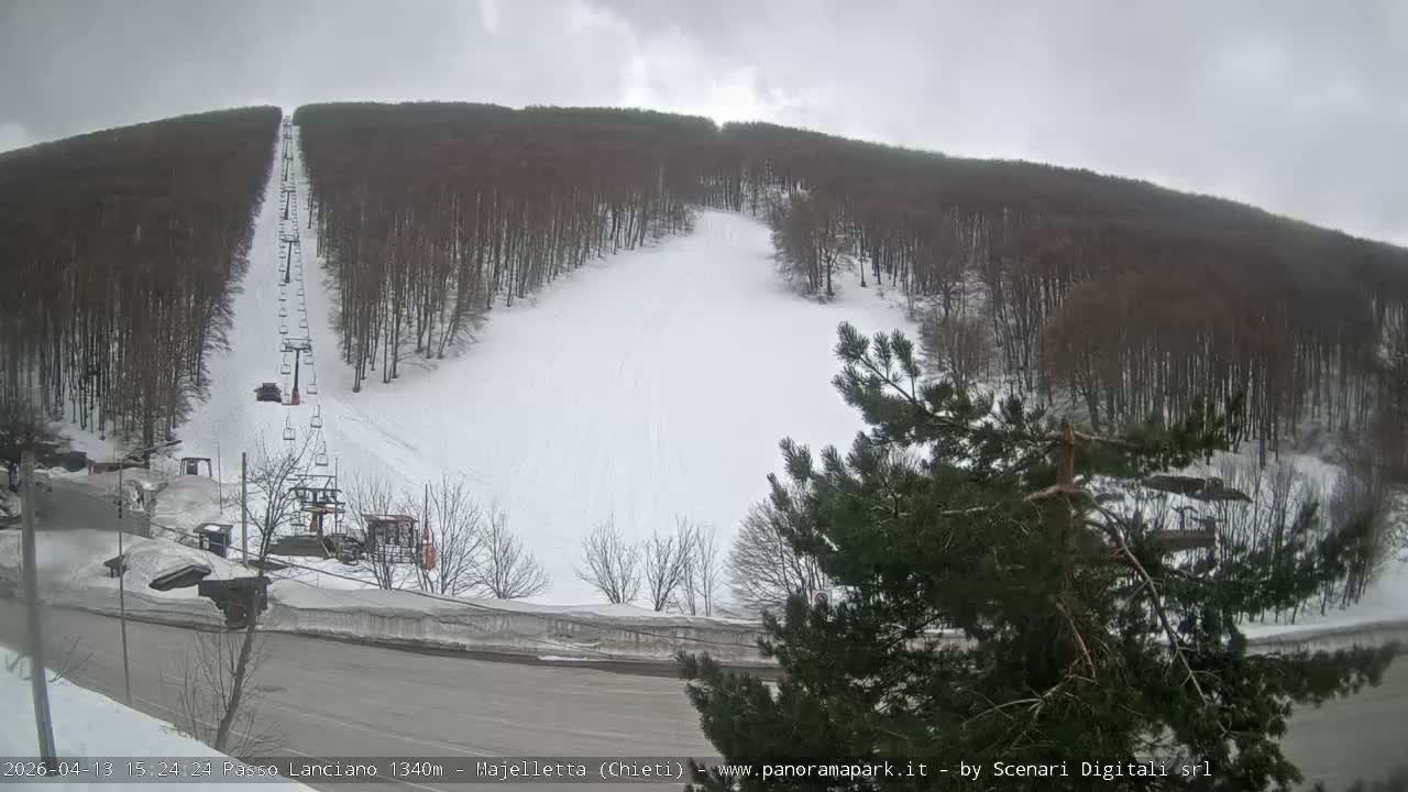 The image captures a snow-covered ski slope with a ski lift ascending between dense forests, a road, and small buildings visible in the foreground, all under an overcast sky.