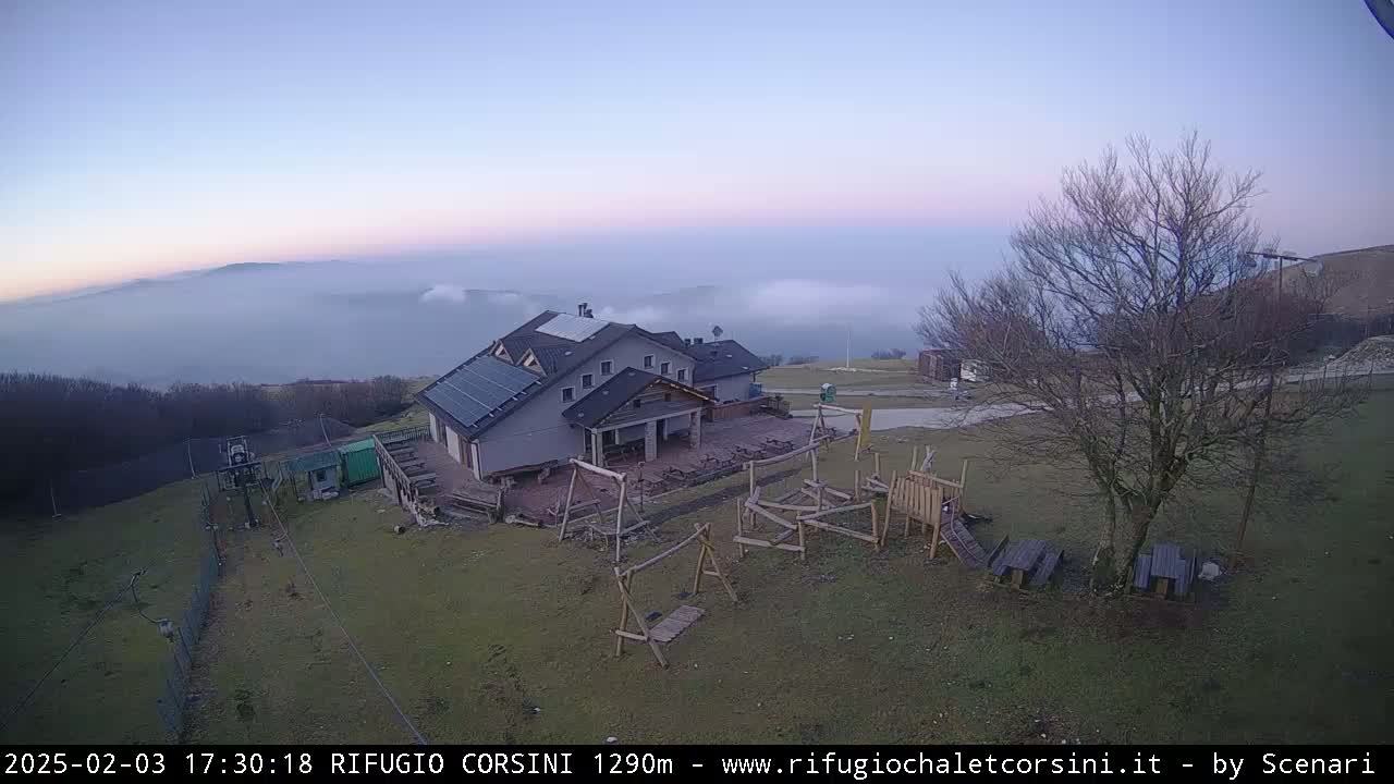 A mountain lodge with a playground sits on a hilltop overlooking a valley filled with fog under a twilight sky.
