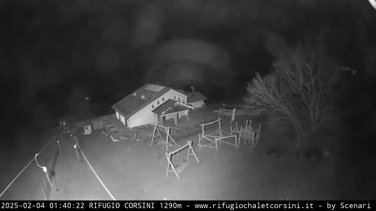 A nighttime, black-and-white view shows a building with a playground, a tree, and what appears to be a small group of people near a fence in the lower left.