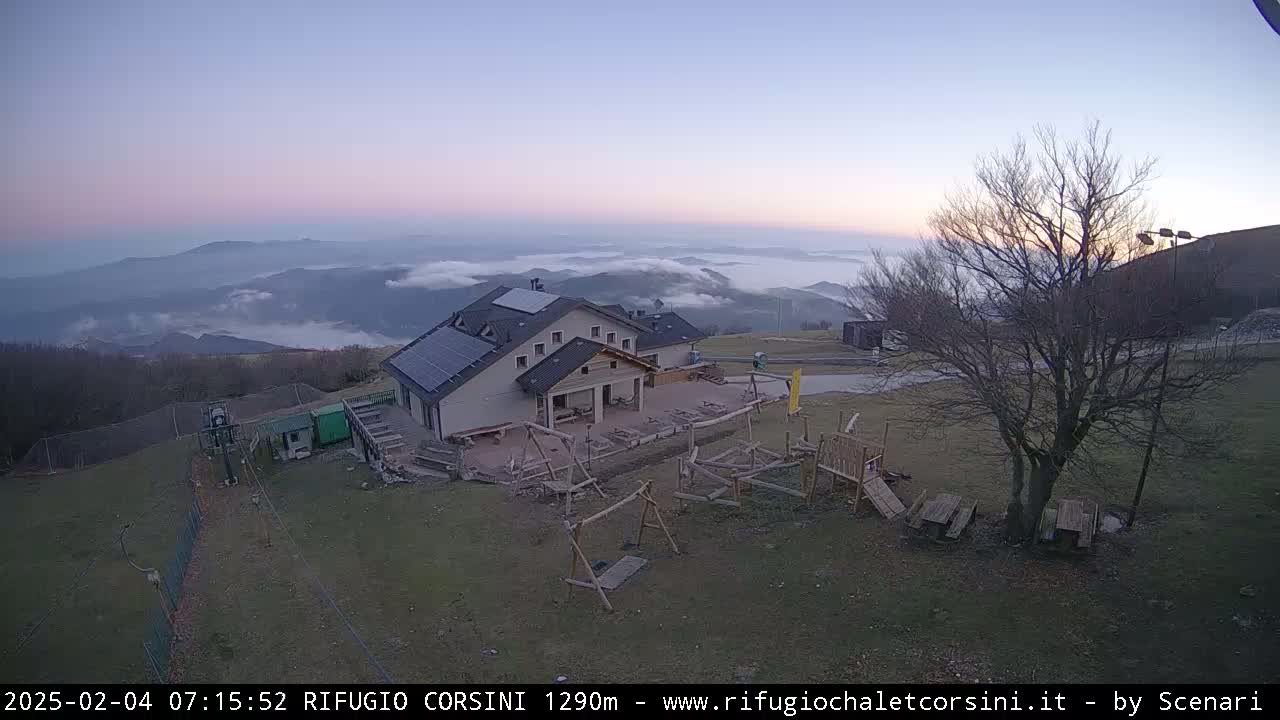 A mountain lodge with a playground sits atop a hill overlooking a valley covered in a layer of fog under a twilight sky.