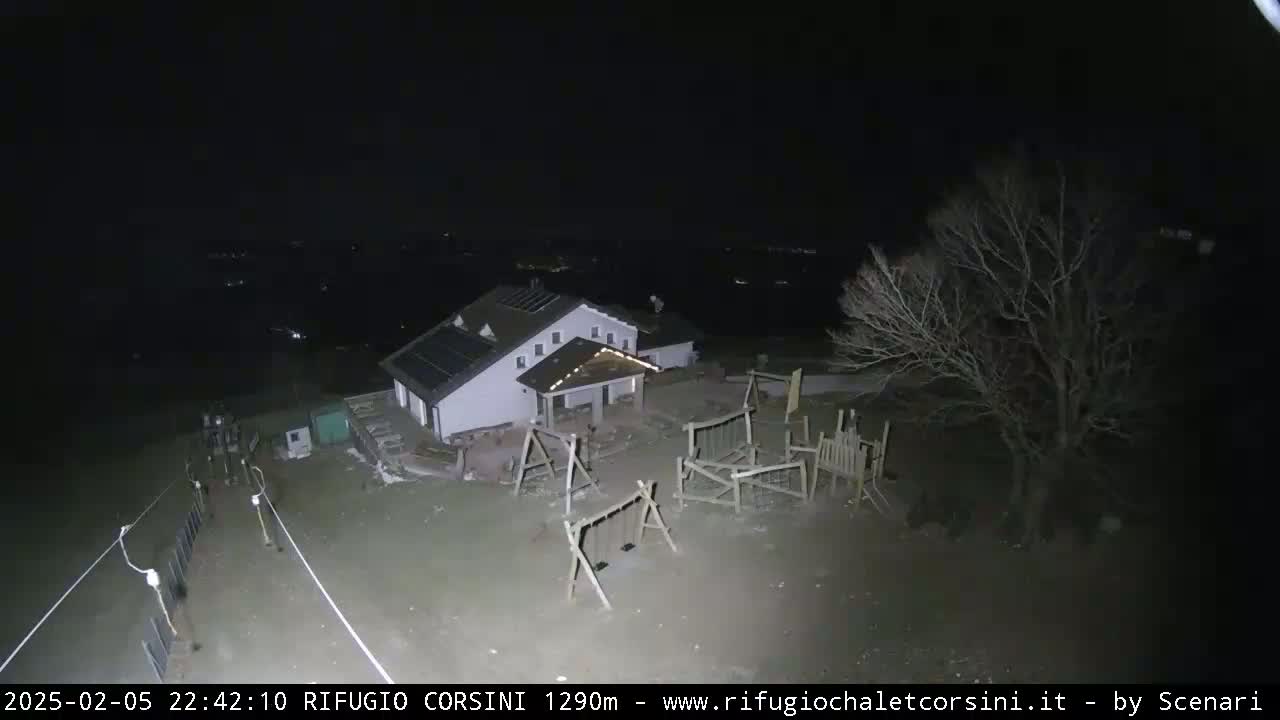A nighttime aerial view shows a building with solar panels on its roof, playground equipment, and a bare tree in a mostly empty, dark yard.