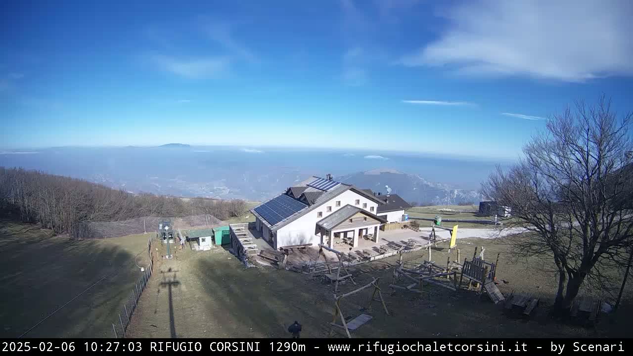 A mountaintop building with solar panels is visible, overlooking a hazy valley under a mostly sunny sky.