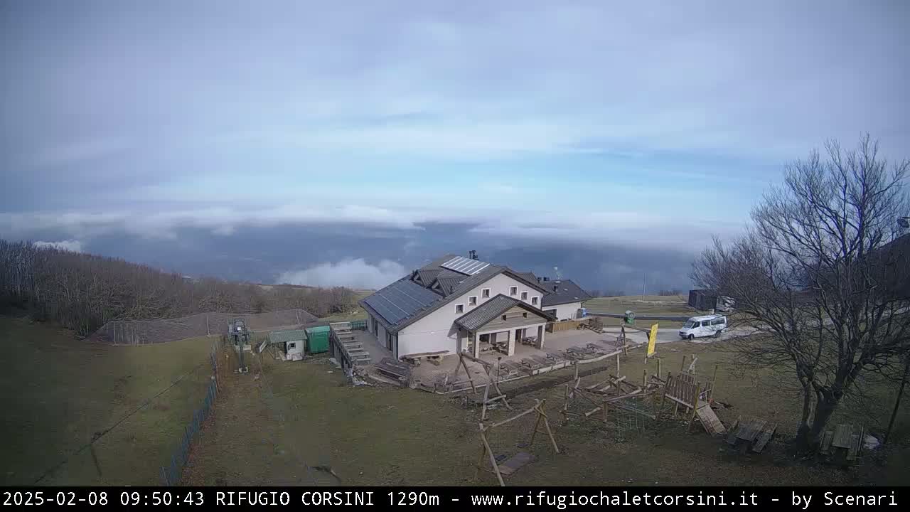 A mountain lodge with solar panels on its roof sits on a grassy area overlooking a valley filled with low-lying clouds under a partly cloudy sky.
