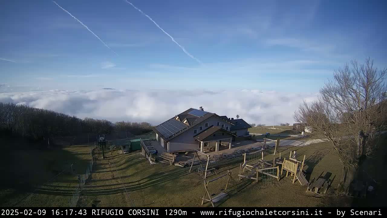 A mountain lodge with solar panels sits atop a hill surrounded by a playground and bare trees, overlooking a sea of clouds under a clear blue sky.