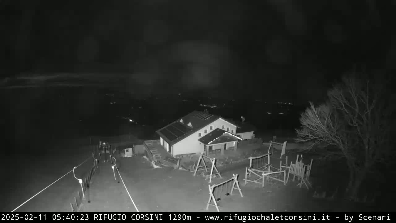 A nighttime, low-light view shows a building with a playground next to it, a sparsely populated area visible in the distance under a dark, cloudy sky.