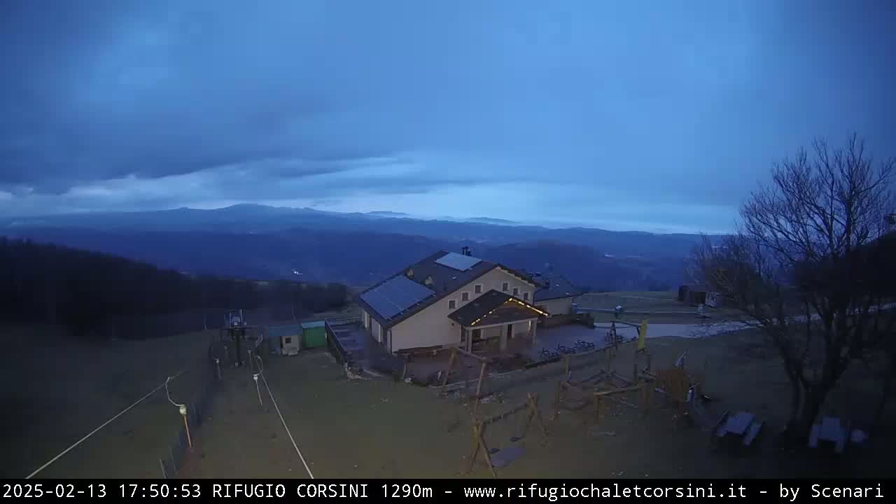 A mountain lodge with solar panels sits on a hill overlooking a valley under a twilight sky.