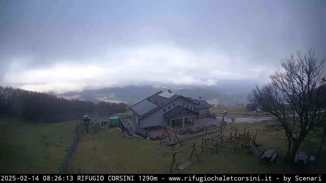 A mountain lodge with a playground sits on a grassy hill under an overcast sky, with a valley shrouded in fog visible in the distance.