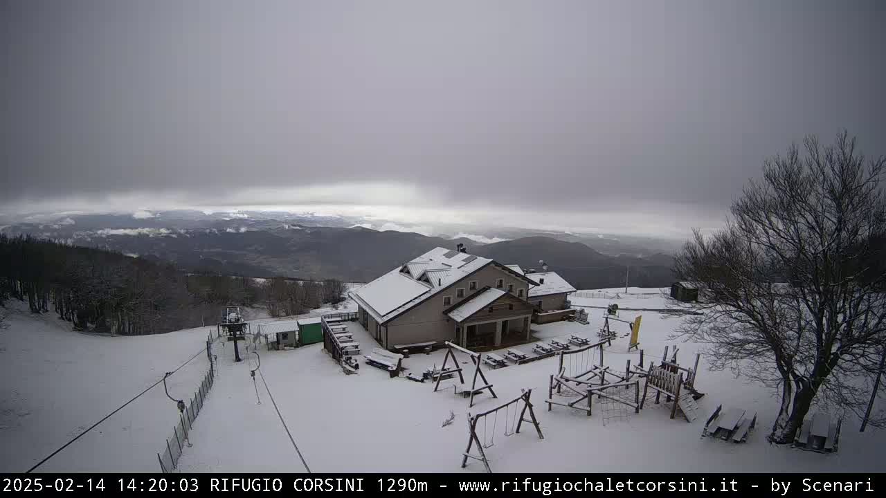 A snow-covered mountain lodge with a playground sits atop a snowy hill overlooking a valley under a cloudy sky.