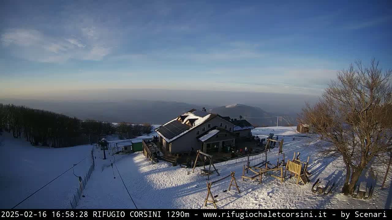 A snow-covered mountaintop scene with a building, playground equipment, and a ski lift under a mostly clear, bright sky.