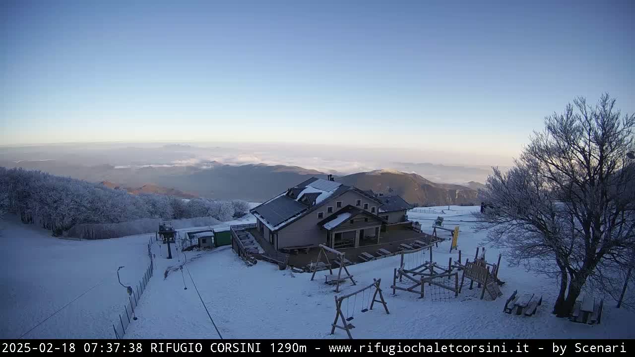 A snow-covered mountaintop lodge with a playground and ski lift, under a clear blue sky with a layer of fog in the distance.