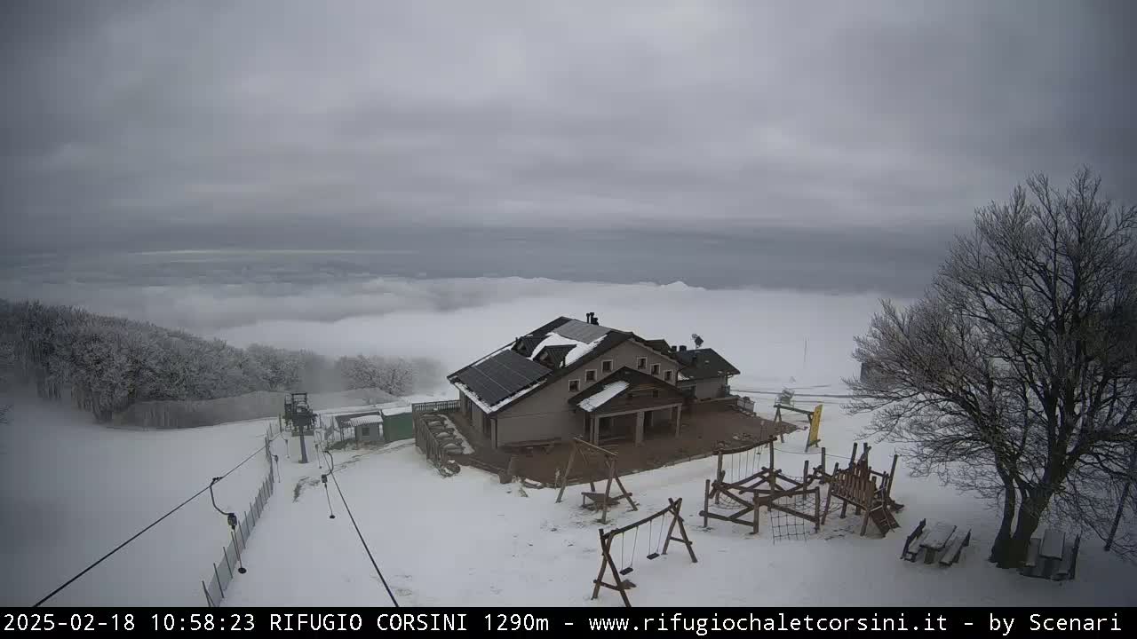 A snow-covered mountain lodge with a playground sits atop a hill shrouded in low-lying clouds under an overcast sky.