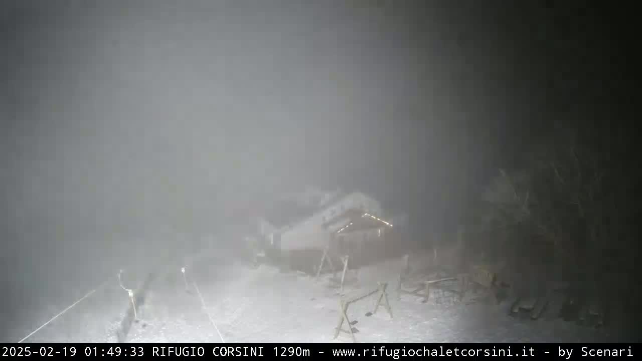 A snow-covered building and outdoor furniture are barely visible through heavy fog at night.