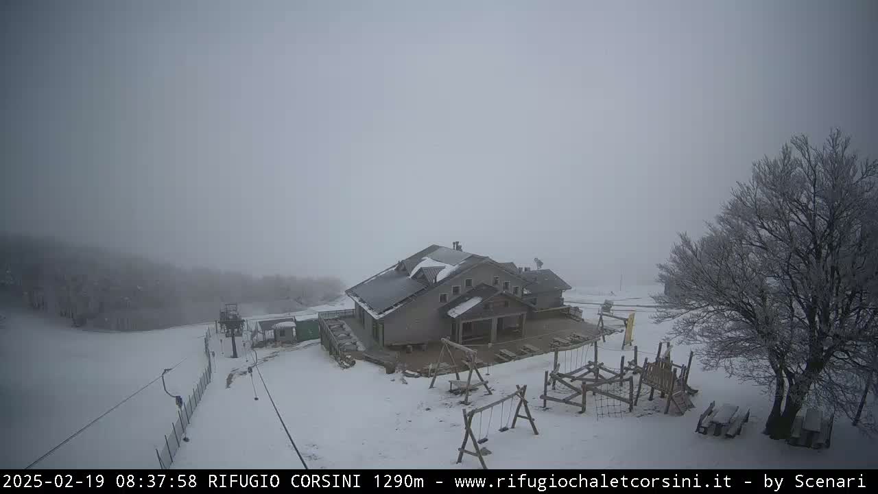 A snow-covered mountaintop scene in foggy conditions shows a building with a playground and a ski lift, and a bare, snow-covered tree.