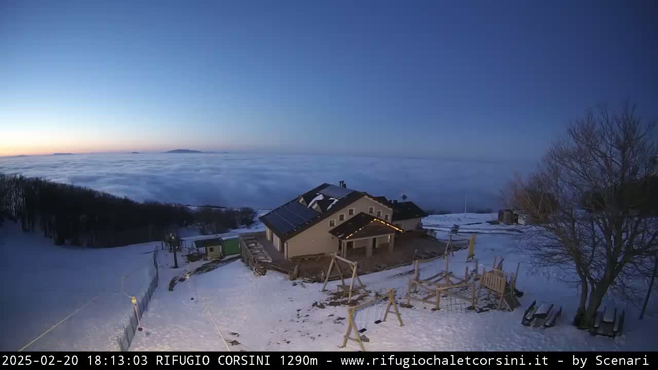 A snow-covered mountaintop building with a playground is situated above a thick layer of clouds under a twilight sky.