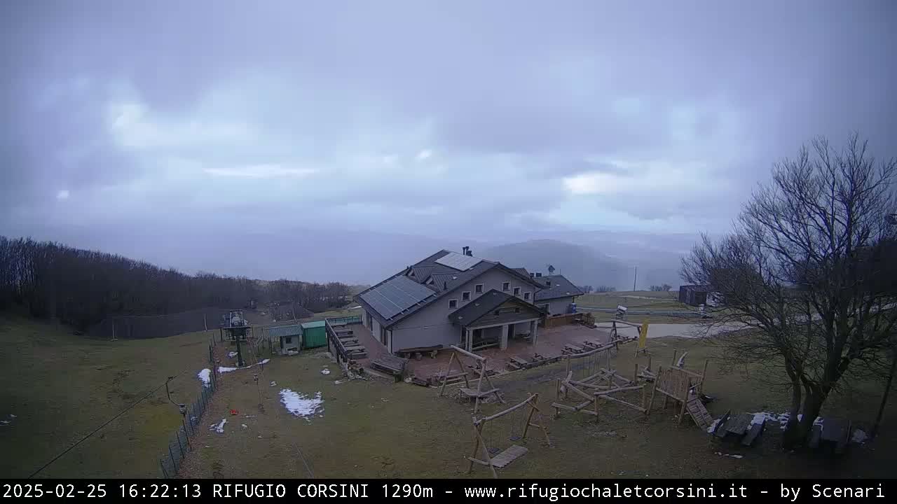 A mountain lodge with solar panels on its roof is situated on a grassy hillside under an overcast sky.