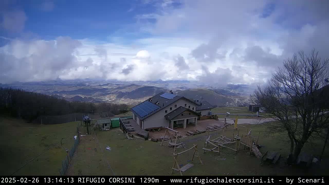 A mountaintop building with solar panels is surrounded by a playground and overlooks a partly cloudy, hilly landscape.