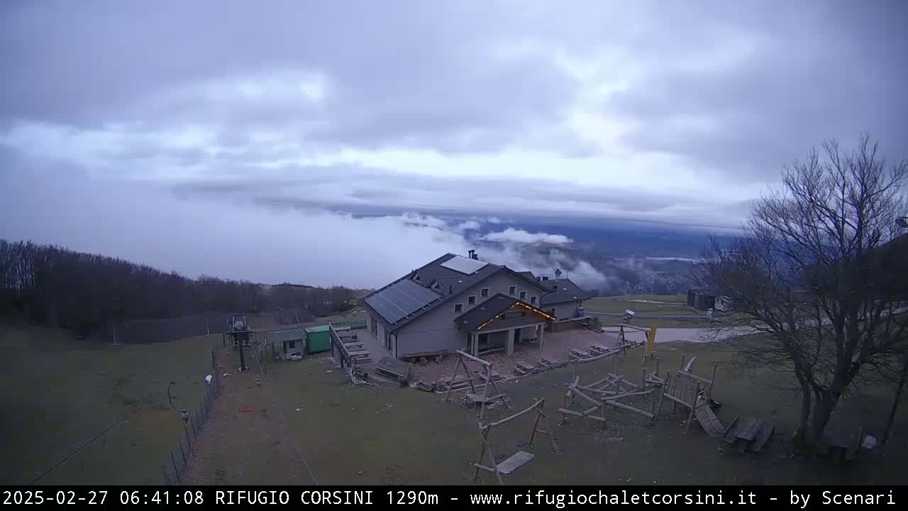 A mountain lodge with solar panels on its roof sits in a clearing surrounded by bare trees and a playground, overlooking a valley shrouded in low-lying clouds under an overcast sky.