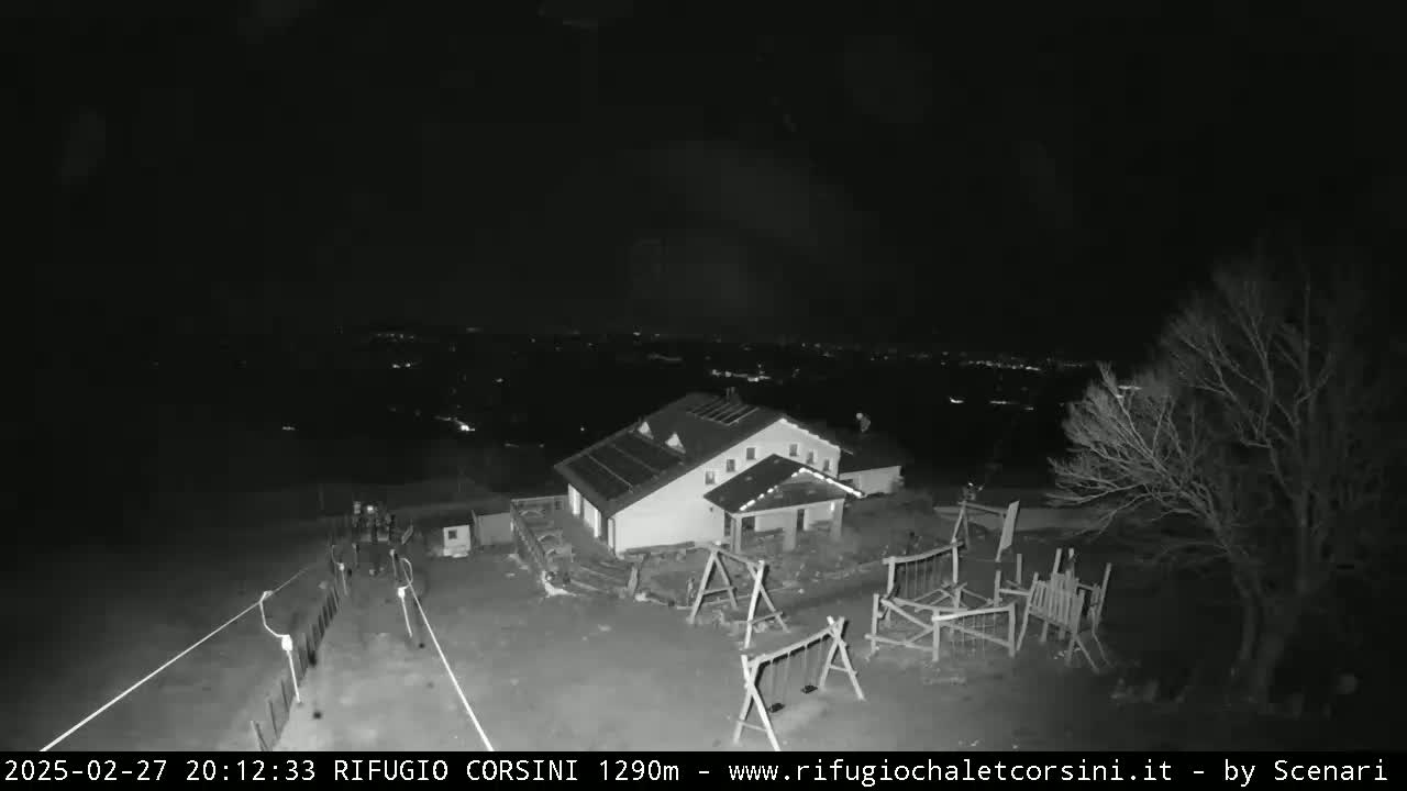 A nighttime, black and white image shows a building with a playground and a few people walking nearby, overlooking a distant city under a clear, dark sky.