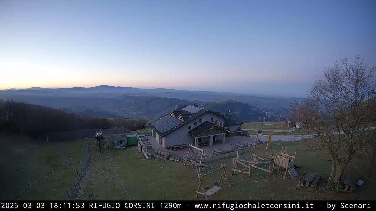 A mountain lodge with solar panels sits on a grassy area overlooking a vast, hazy valley at dusk.