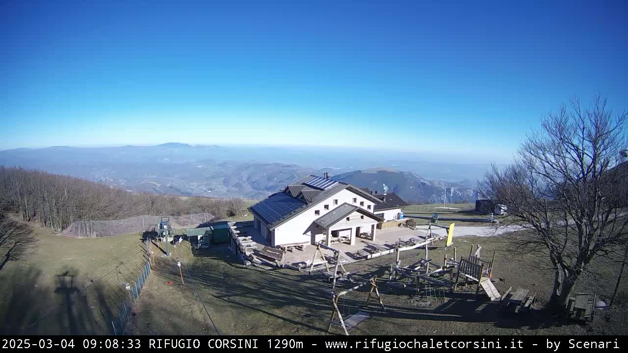 A mountaintop building with solar panels sits next to a playground, overlooking a vast valley under a clear blue sky.