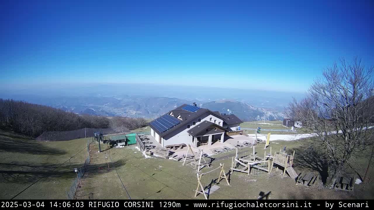 A mountaintop building with solar panels sits below a clear blue sky, overlooking a valley and featuring a playground and ski lift in the foreground.