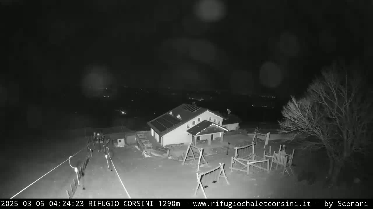 A nighttime, black and white overhead view shows a building with solar panels, a playground, and some bare trees under a dark, possibly cloudy sky.