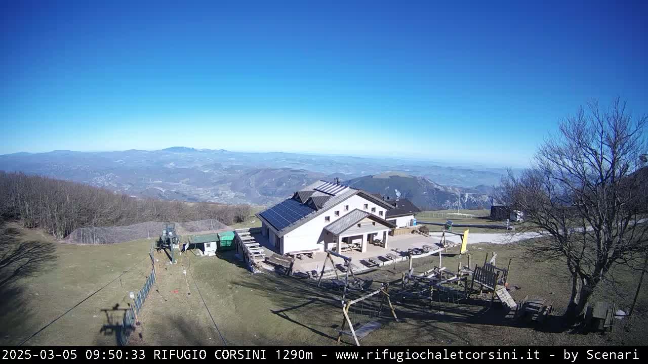 An aerial view shows a building with solar panels on its roof, playground equipment, and a ski lift in a mountainous area under clear, sunny skies.