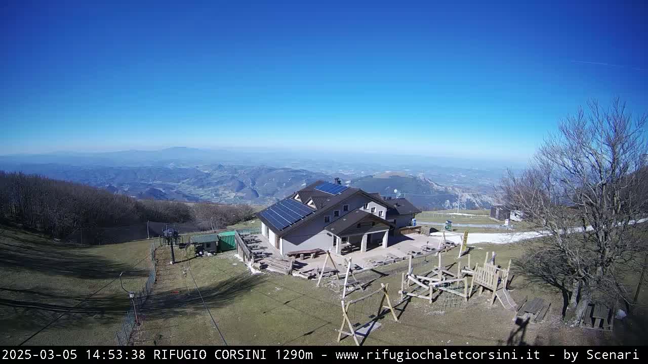 A mountaintop building with solar panels, a playground, and a ski lift is viewed on a clear, sunny day with a vast valley visible in the background.
