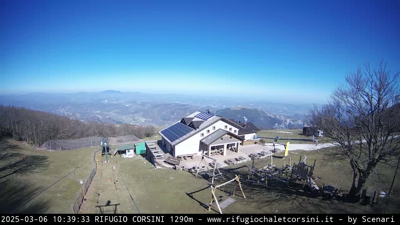 A mountaintop building with solar panels, a playground, and a ski lift, overlooks a valley on a clear, sunny day.