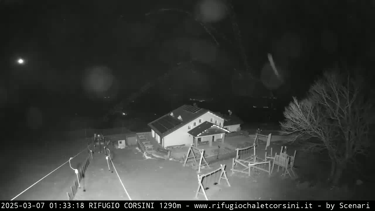 A nighttime, snowy scene shows a building with solar panels, a playground, and several people walking near a road.
