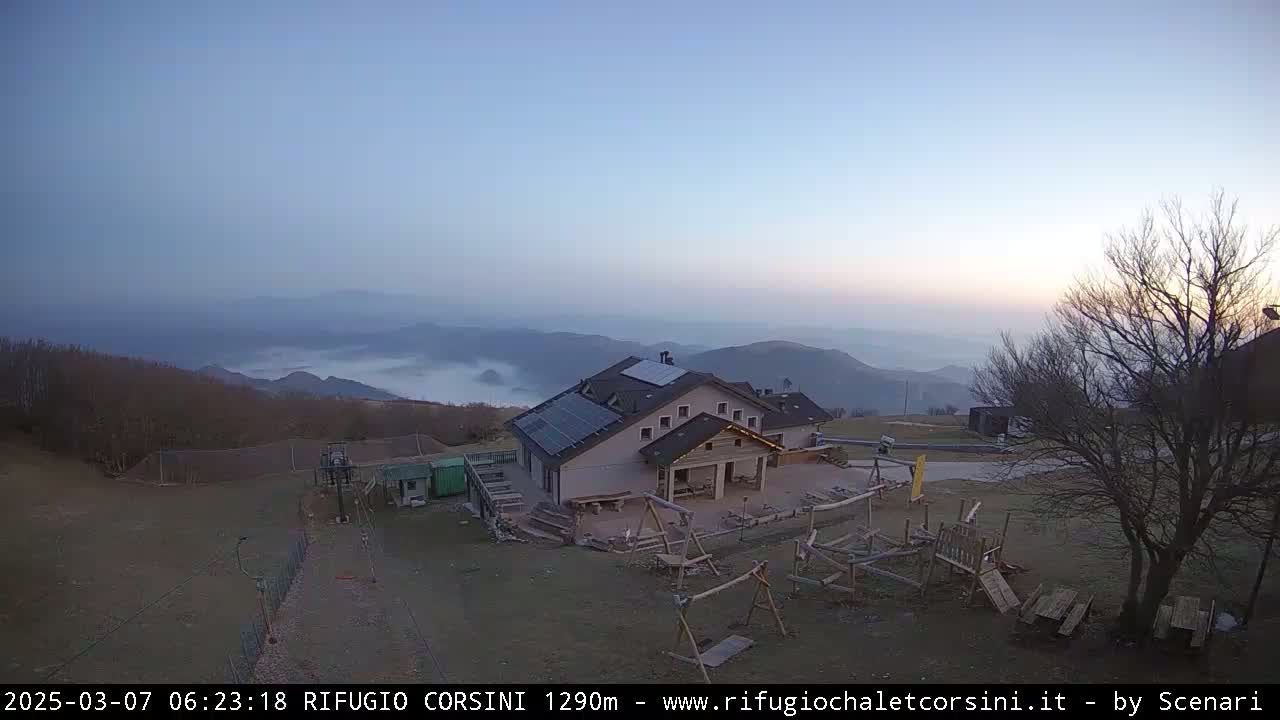 A mountaintop building with solar panels is visible, overlooking a valley filled with low-lying fog under a clear, pale sky.
