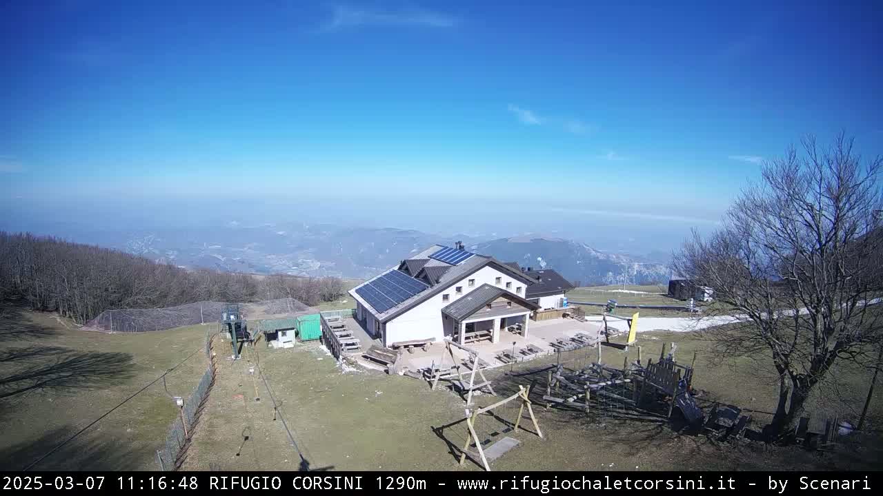 A mountaintop building with solar panels sits on a grassy area with a playground, overlooking a hazy valley under a clear blue sky.
