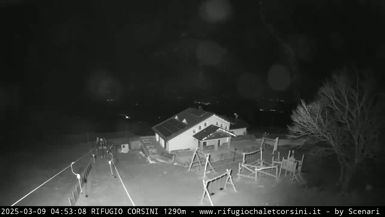 A nighttime, black and white view of a building with a playground and a few people near it, under a dark, cloudy sky.