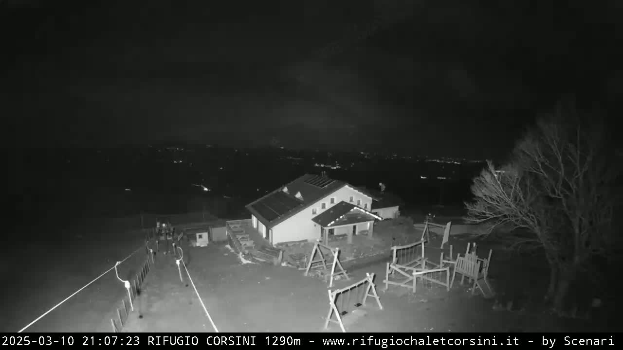 A nighttime, low-light view shows a building with a playground and surrounding area, overlooking a distant, dimly lit town.