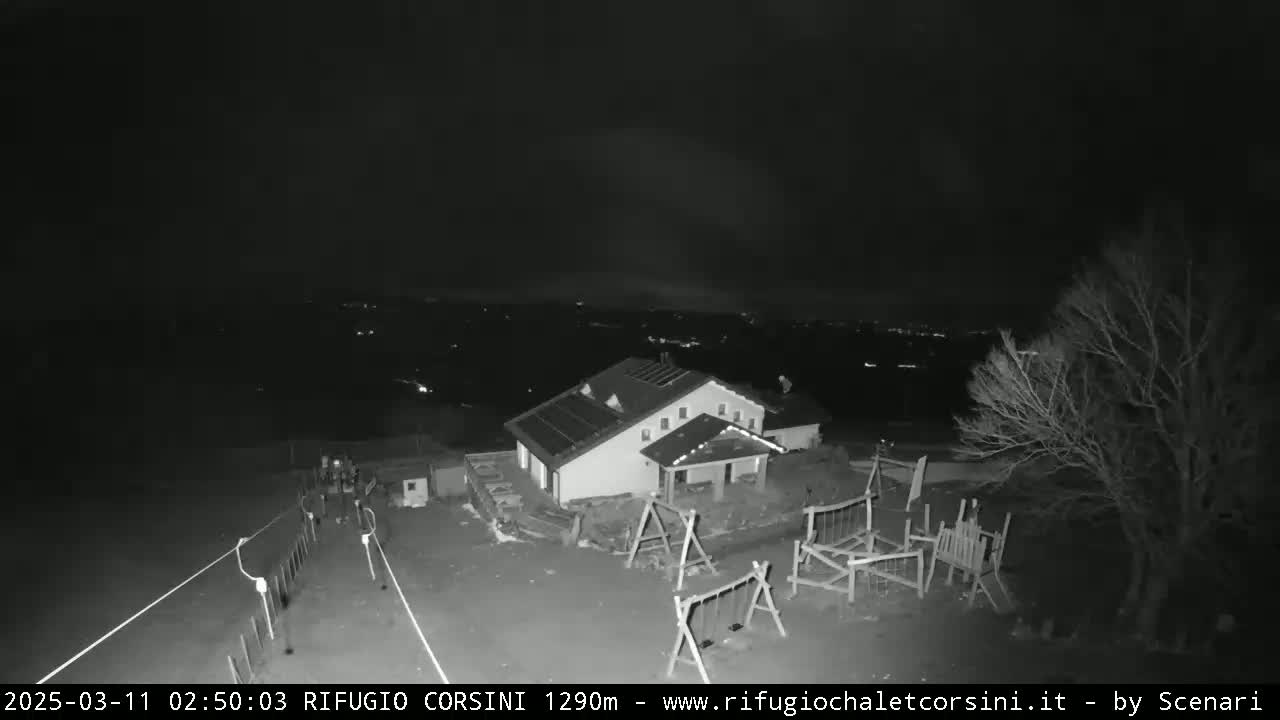 A nighttime, black and white image shows a building with a playground and some people near a fence, overlooking a distant, dimly lit landscape under a dark sky.