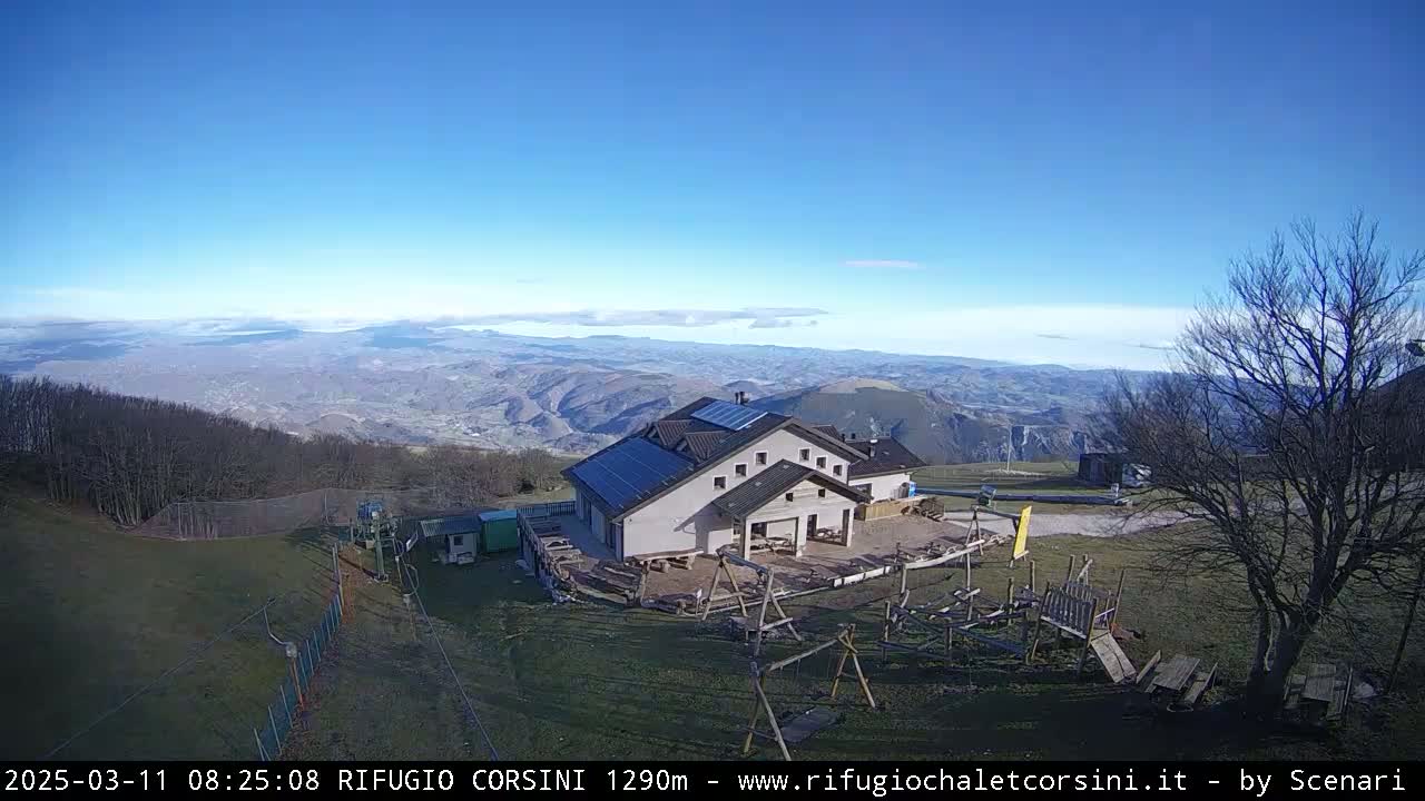 A mountaintop building with solar panels is shown, overlooking a vast valley under a clear, sunny sky.