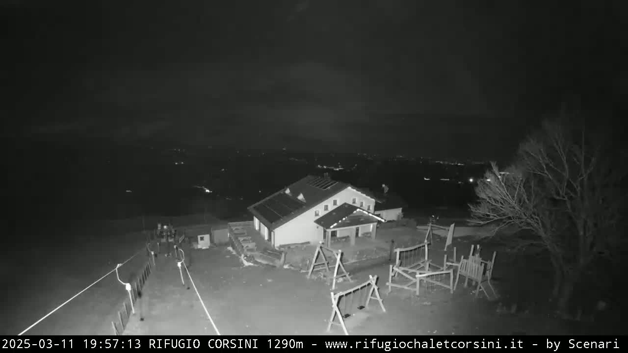 A nighttime, low-light image shows a building with solar panels on its roof, a playground, and a bare tree, all situated on a hill overlooking a distant, dimly lit town.
