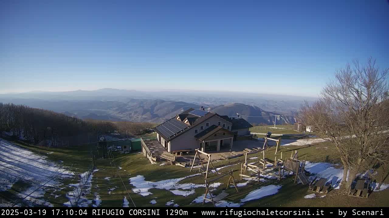 A mountaintop building with solar panels is seen, overlooking a valley with patches of snow under a clear blue sky.