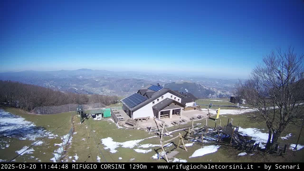 A mountaintop building with solar panels is visible, overlooking a vast valley on a clear, sunny day with patches of snow on the ground.